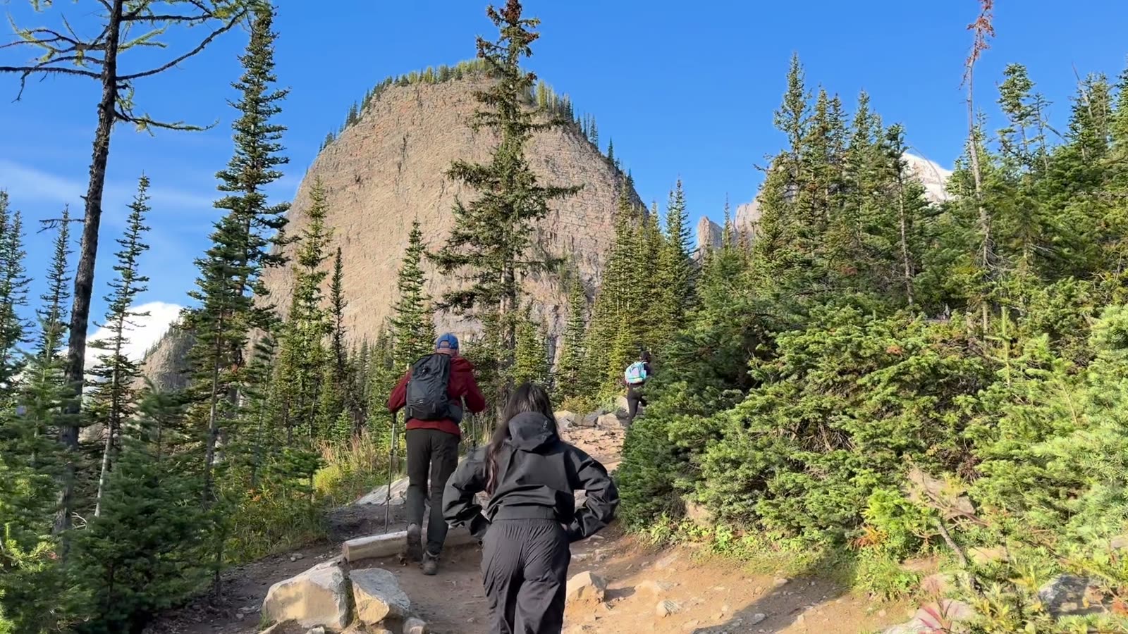 Three hikers walking up a forest trail toward a rounded rock peak, shown over the line “choosing my three columns and pillars of my life and then working towards that.”
